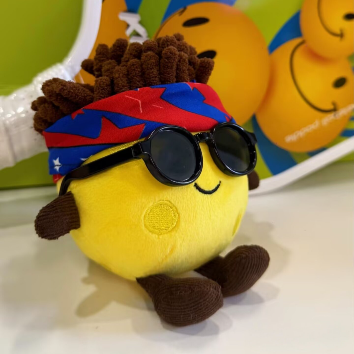 Yellow plush toy with sunglasses and a colorful headband on a white surface.