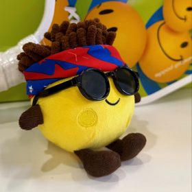 Yellow plush toy with sunglasses and a colorful headband on a white surface.