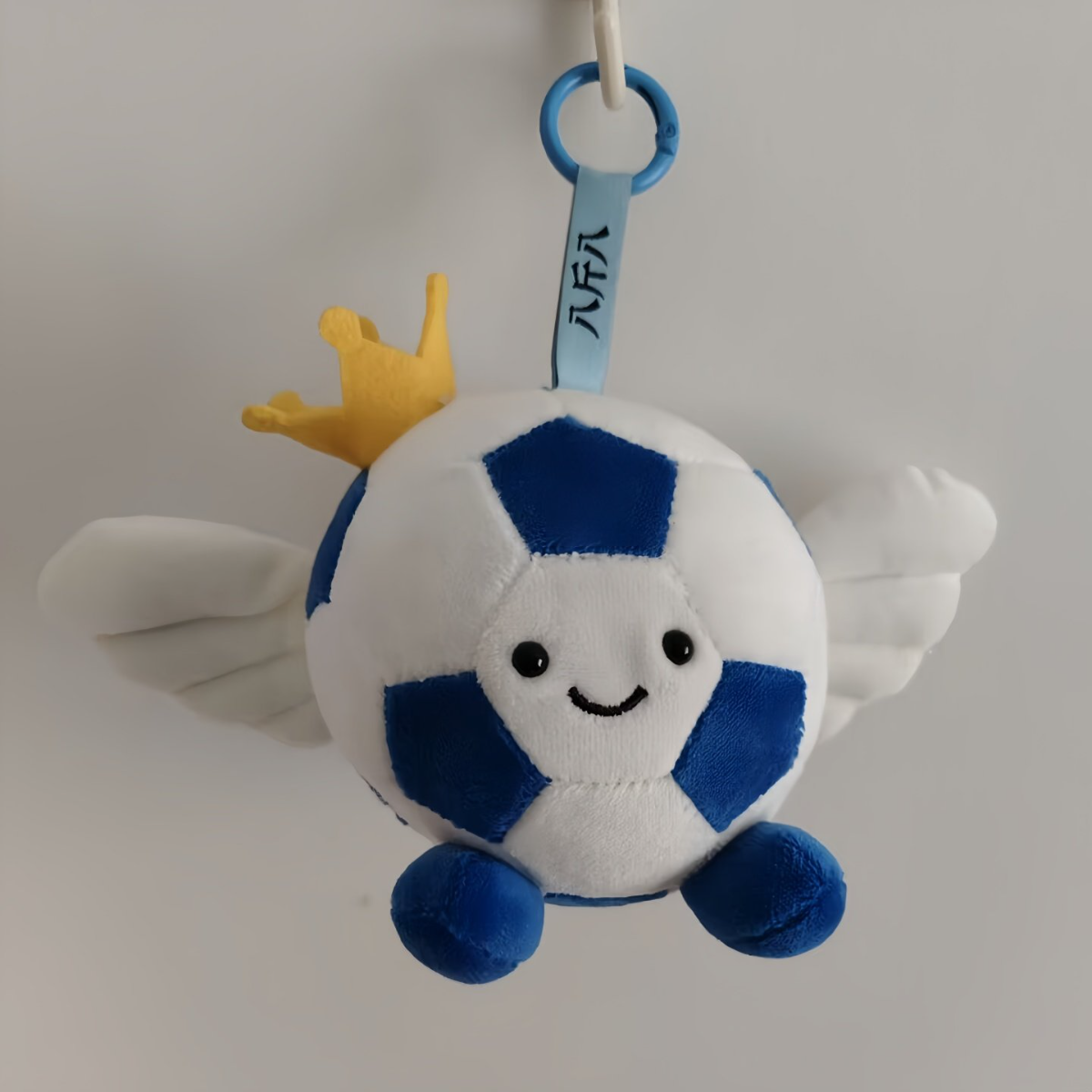 Plush toy resembling a soccer ball with wings, a crown, and a tag on a plain background